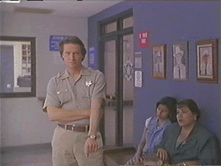 Chris Cooper as Sam Deeds stands, grabbing his left elbow with his right hand